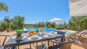 Un área de comedor al aire libre con piscina al fondo, rodeada de follaje.