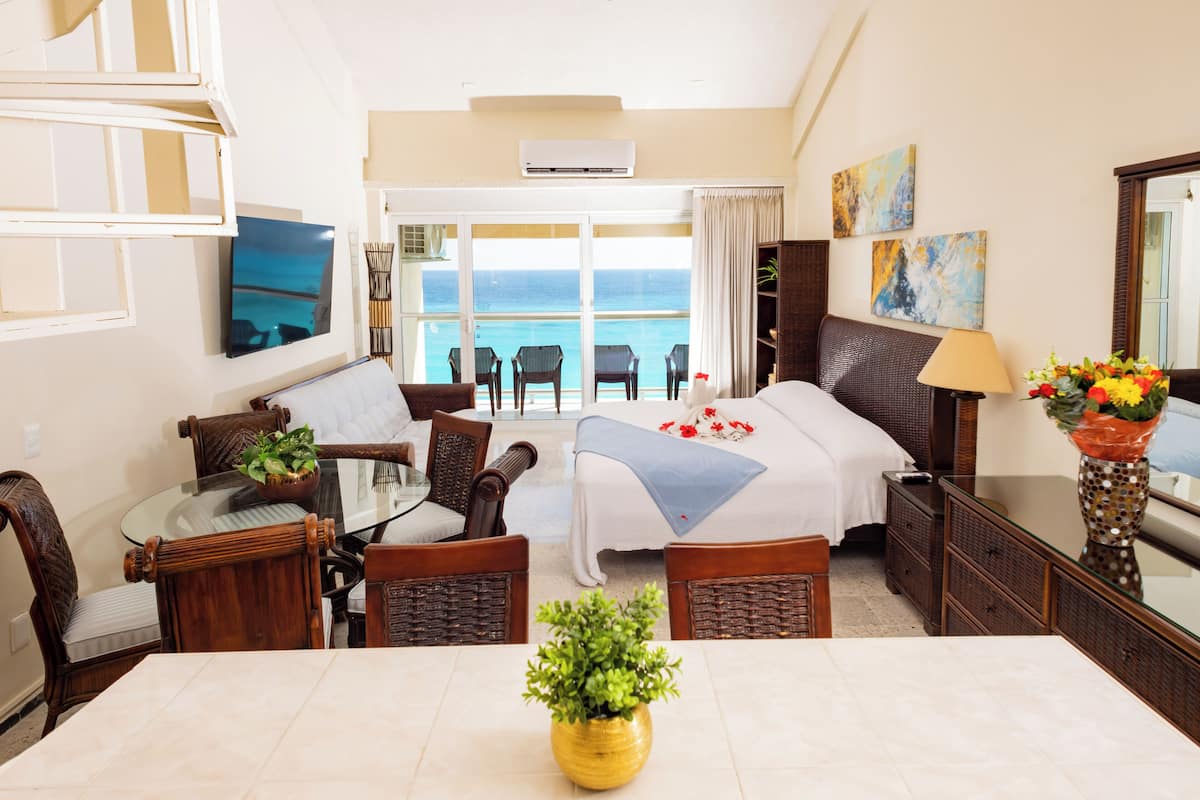 Une chambre d’hôtel comprenant un lit, une table à manger avec des chaises, une télévision, un balcon avec vue sur la mer et un vase rempli de fleurs.