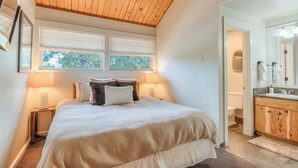 Une chambre à coucher comprenant un lit, des lampes de chevet, un plafond en bois, une fenêtre et une salle de bain avec un lavabo et un miroir.