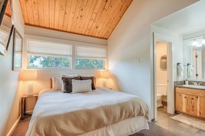 A bedroom with a bed, bedside lamps, a wooden ceiling, a window, and a bathroom with a sink and mirror.