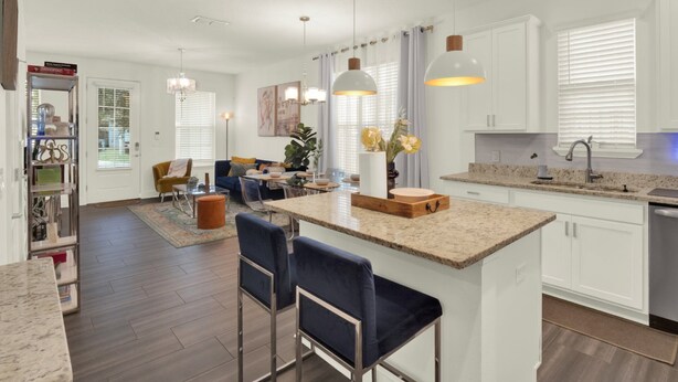 A modern kitchen with a central island, bar stools, pendant lighting, and a dining area with a sofa and coffee table.