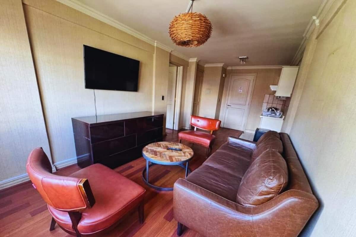 Ein Wohnzimmer mit braunem Ledersofa, einem roten Ledersessel und einem runden Couchtisch. Ein Flachbildfernseher ist an der Wand montiert, und eine Weidenlampe hängt von der Decke.