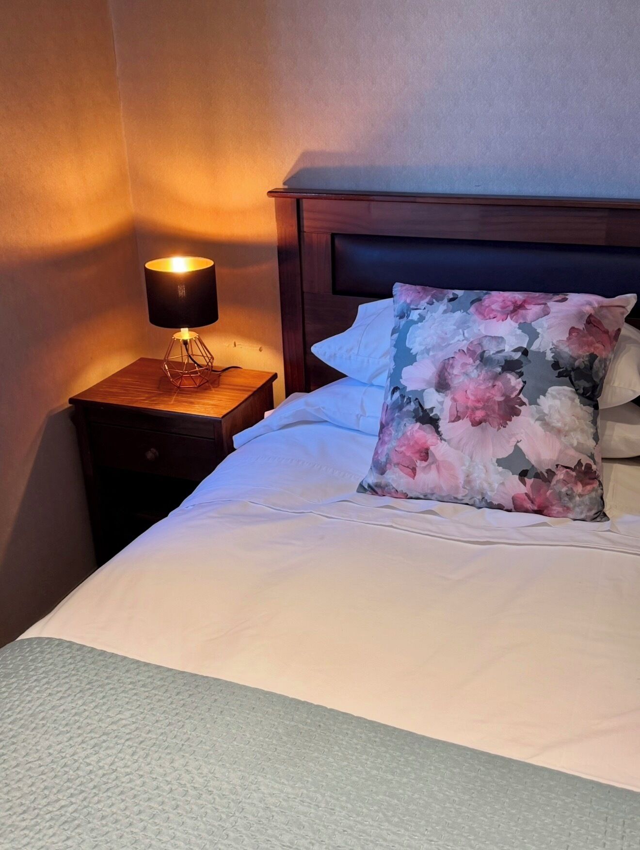 Un lit bien fait, agrémenté d’un coussin à motifs floraux, d’une lampe de chevet et d’une table de nuit en bois.