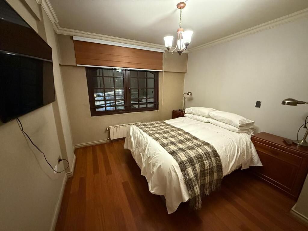 Un dormitorio con cama, mesita de noche, lámpara, radiador, ventana con persianas y televisor montado en la pared.