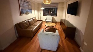 Ein Wohnzimmer mit braunem Ledersofa, einem gemusterten Ottomanen, einem runden Couchtisch, einem weißen Sessel, einer Pendelleuchte und einem flachbildfernseher an der Wand.