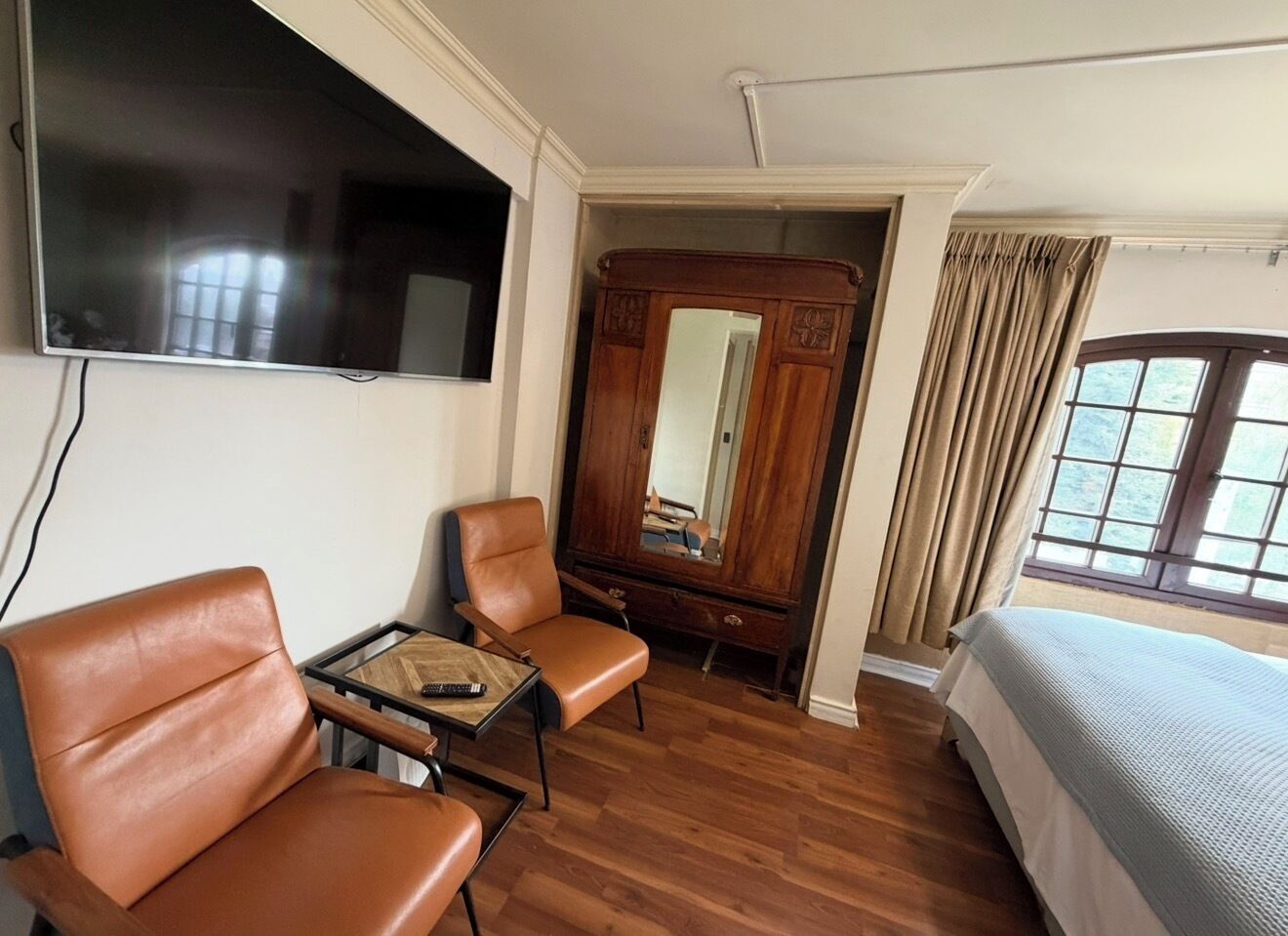 A hotel room with a bed, two leather chairs, a small table with a remote control, a wooden wardrobe, a television mounted on the wall, and a window with curtains.