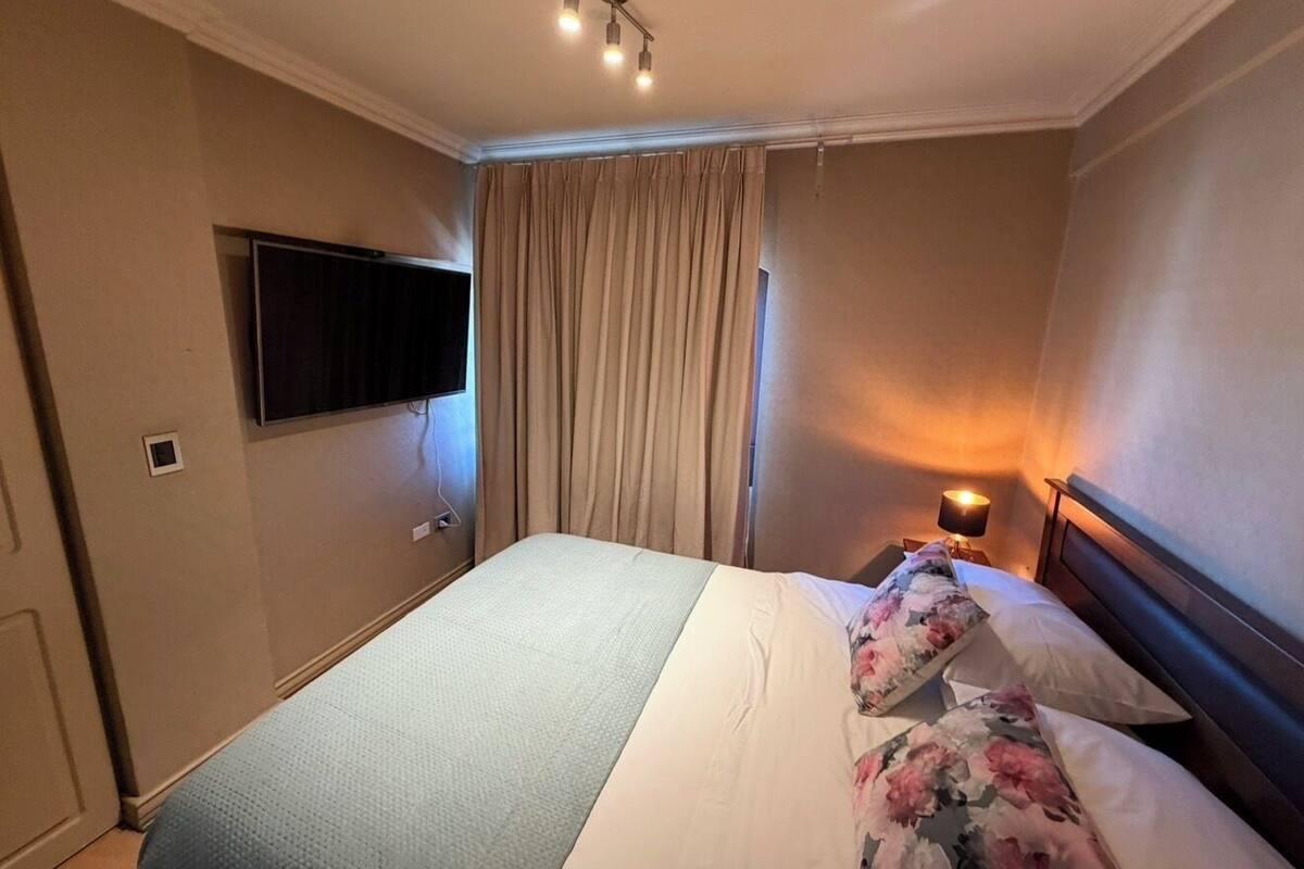 Ein Hotelzimmer mit Bett, Vorhängen, Fernseher, Nachttisch mit Lampe und Wand-Fernseher.
