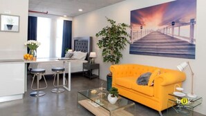 A modern living room with a glass coffee table, a bright orange sofa, and a large painting of a sunset over a pier.