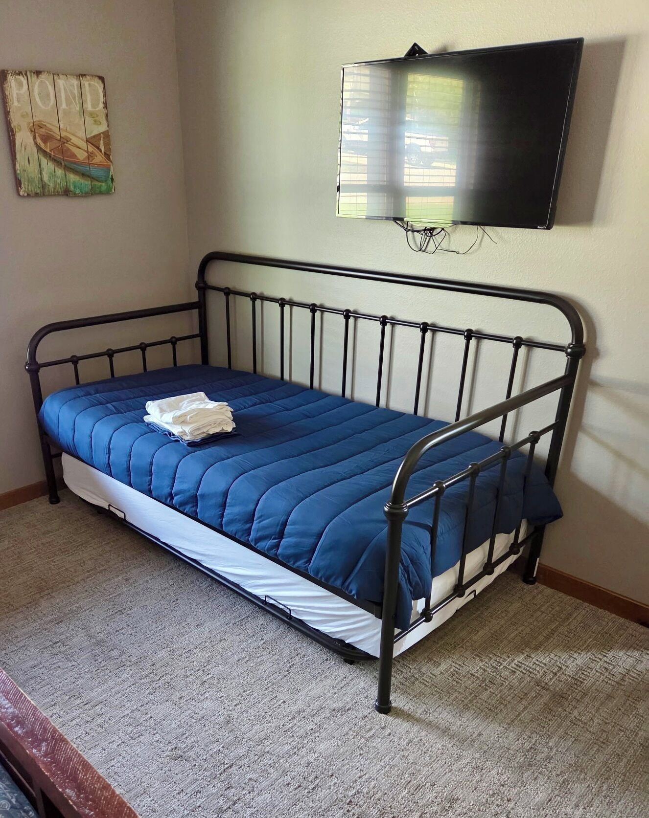 A single bed with a blue mattress and white sheets, a wall-mounted television, and a small painting on the wall.