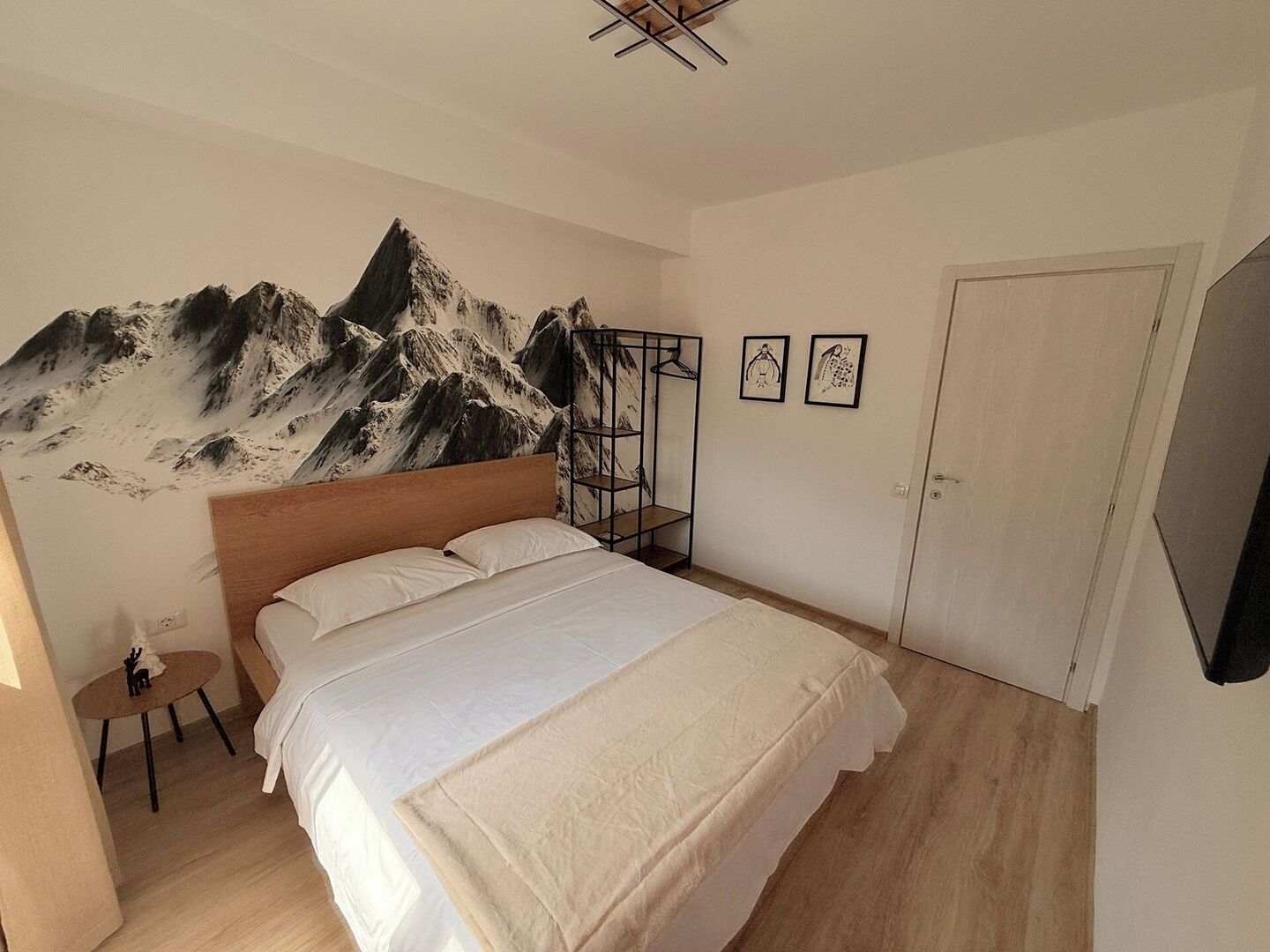 Een slaapkamer met een bed, een houten hoofdbord, een kapstok aan de muur, een klein tafeltje met een stoel, en een deur.