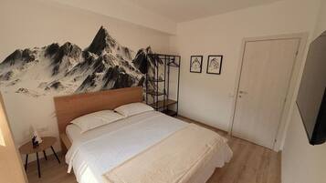 Een slaapkamer met een bed, een houten hoofdbord, een kapstok aan de muur, een klein tafeltje met een stoel, en een deur.