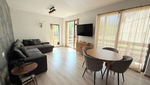 Een moderne woonkamer met een hoekbank, een ronde eettafel met vier stoelen, een flatscreen televisie aan de muur en grote ramen met lichte gordijnen.