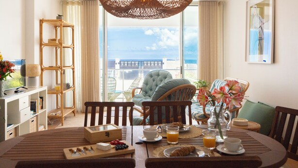 Un coin repas avec vue sur la mer, un lustre en osier et un buffet en bois agrémenté de livres et d’un vase.