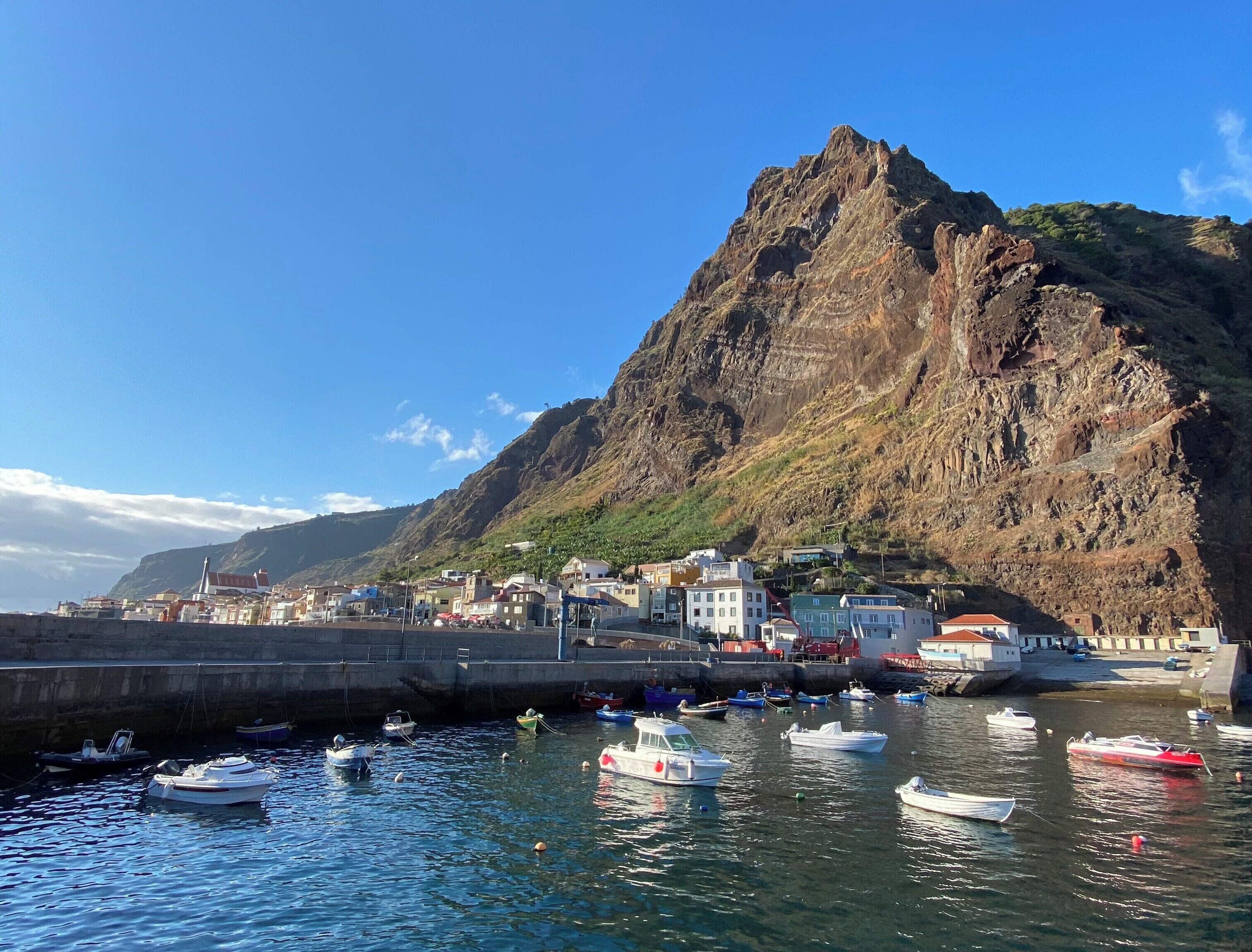Uma vila costeira com uma colina rochosa imponente, um porto com diversos barcos e edifícios ao longo da orla.