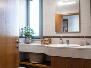 A bathroom with a wooden door, a window, a sink, a mirror, a vase with flowers, and a woven basket.
