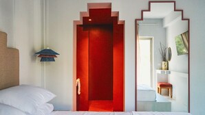 Een moderne hotelkamer met een rode deur, witte muren, een bed met witte lakens, een klein nachtkastje met een lamp, en een spiegel die de kamer weerspiegelt.