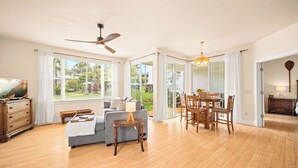 A spacious living room with a sofa, dining table, and chairs, large windows, and a ceiling fan. - Beautiful 2bdrm Condo Facing Lawn, Pool & Hot Tub! (Princeville)