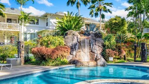 A swimming pool with a waterfall feature, surrounded by lush tropical plants and palm trees. - Beautiful 2bdrm Condo Facing Lawn, Pool & Hot Tub! (Princeville)