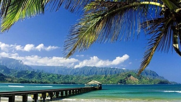 A beach with a wooden pier extending into the ocean, a small hut at the end, and a mountain range in the background. - Beautiful 2bdrm Condo Facing Lawn, Pool & Hot Tub! (Princeville)