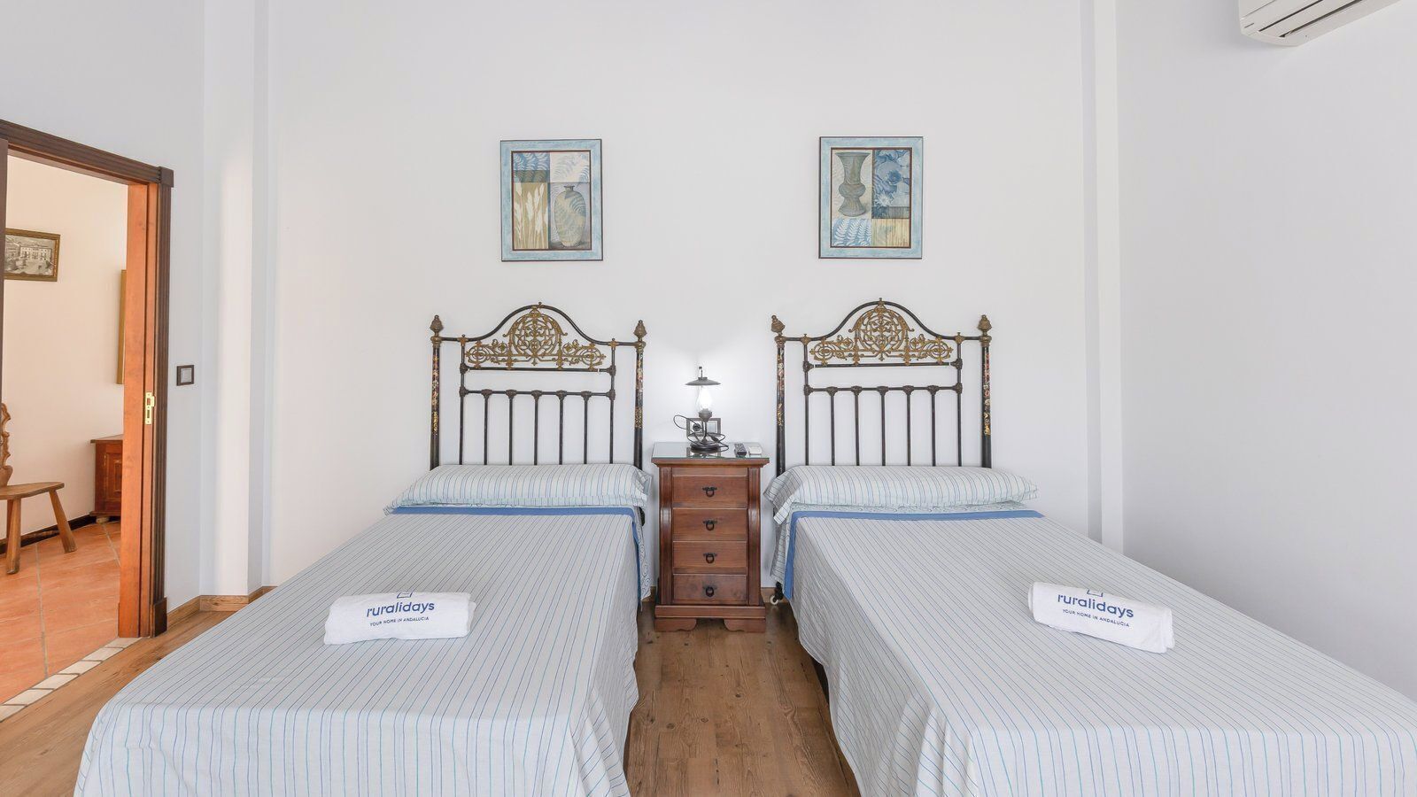 A room with two single beds, a wooden nightstand, a lamp, and two framed pictures on the wall.