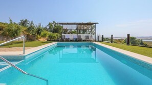 A rectangular swimming pool with clear blue water, surrounded by a grassy area and a covered seating area with chairs and umbrellas.