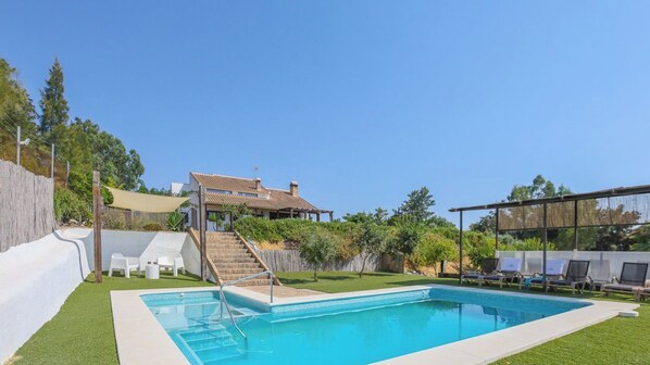 A swimming pool with steps leading to a house, surrounded by greenery and a clear sky.