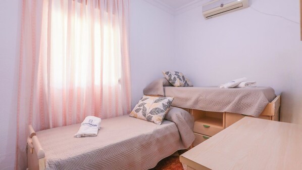 A small, neatly arranged room with two single beds, a wooden table, and a window with sheer curtains.