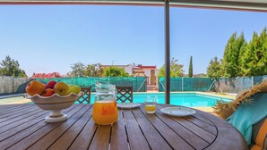 A wooden table with a fruit bowl, a pitcher of orange juice, and a glass of water, set against a backdrop of a swimming pool and a house.