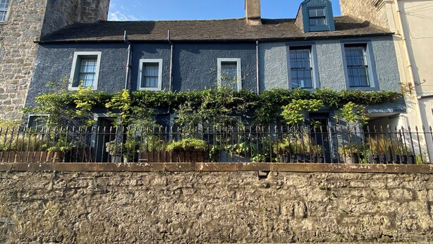 A stone wall with a metal fence in front of a building with multiple windows and a stone chimney.