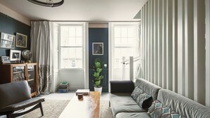 A living room with a sofa, a coffee table, a bookshelf, and a large window with curtains.
