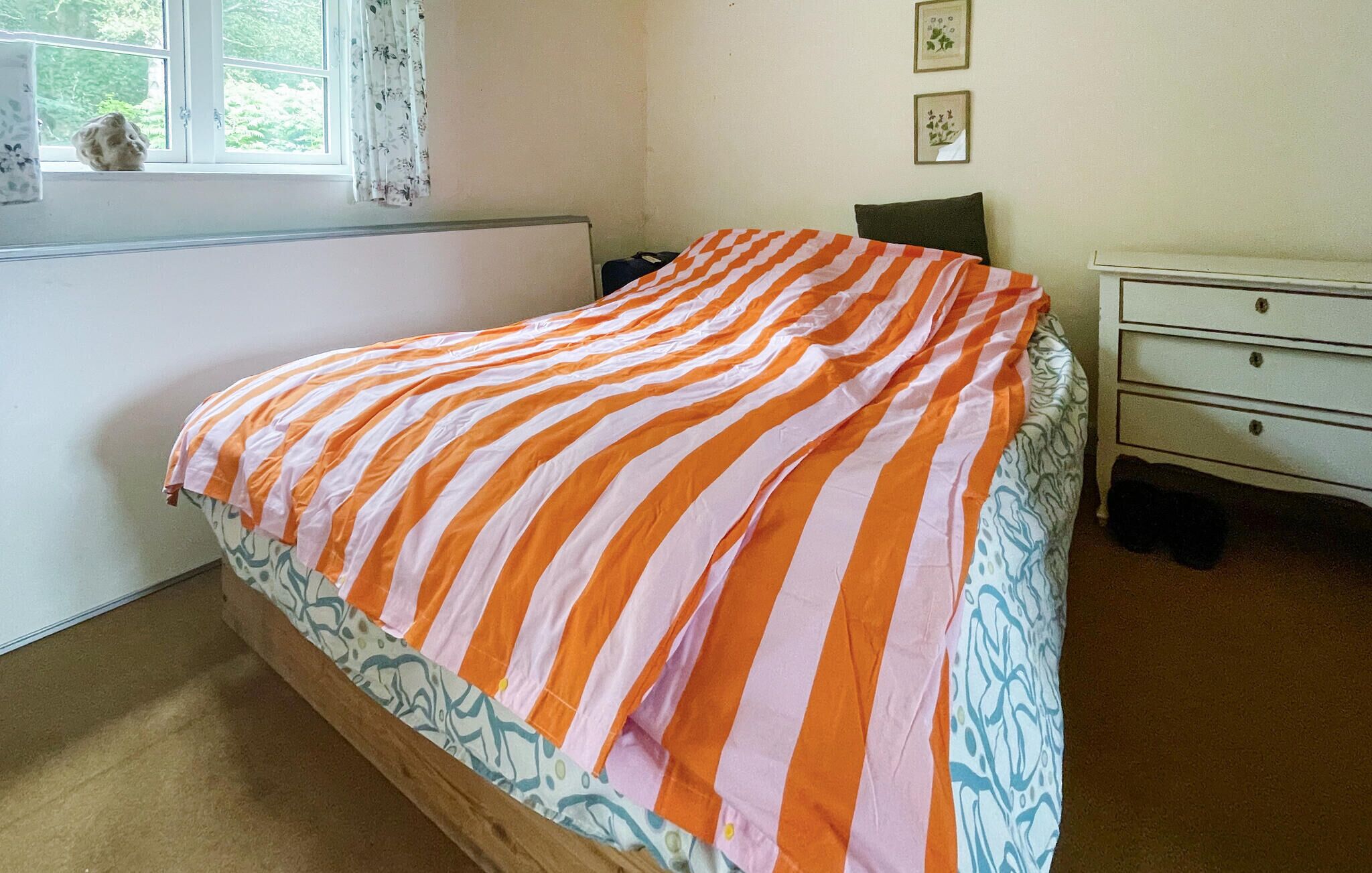 Ein Schlafzimmer mit einem Bett, das mit einer gestreiften Decke bezogen ist, einem Fenster mit Vorhängen, einem Nachttisch und einer Kommode.