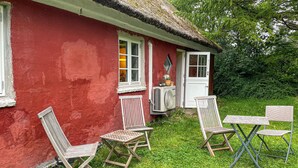 A red house with a thatched roof, a white door, and a window with a white frame. There are two wooden chairs and a small table outside.