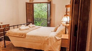 A neatly made bed with a beige bedspread and pillows, a bedside lamp, and a view of greenery through an open door.
