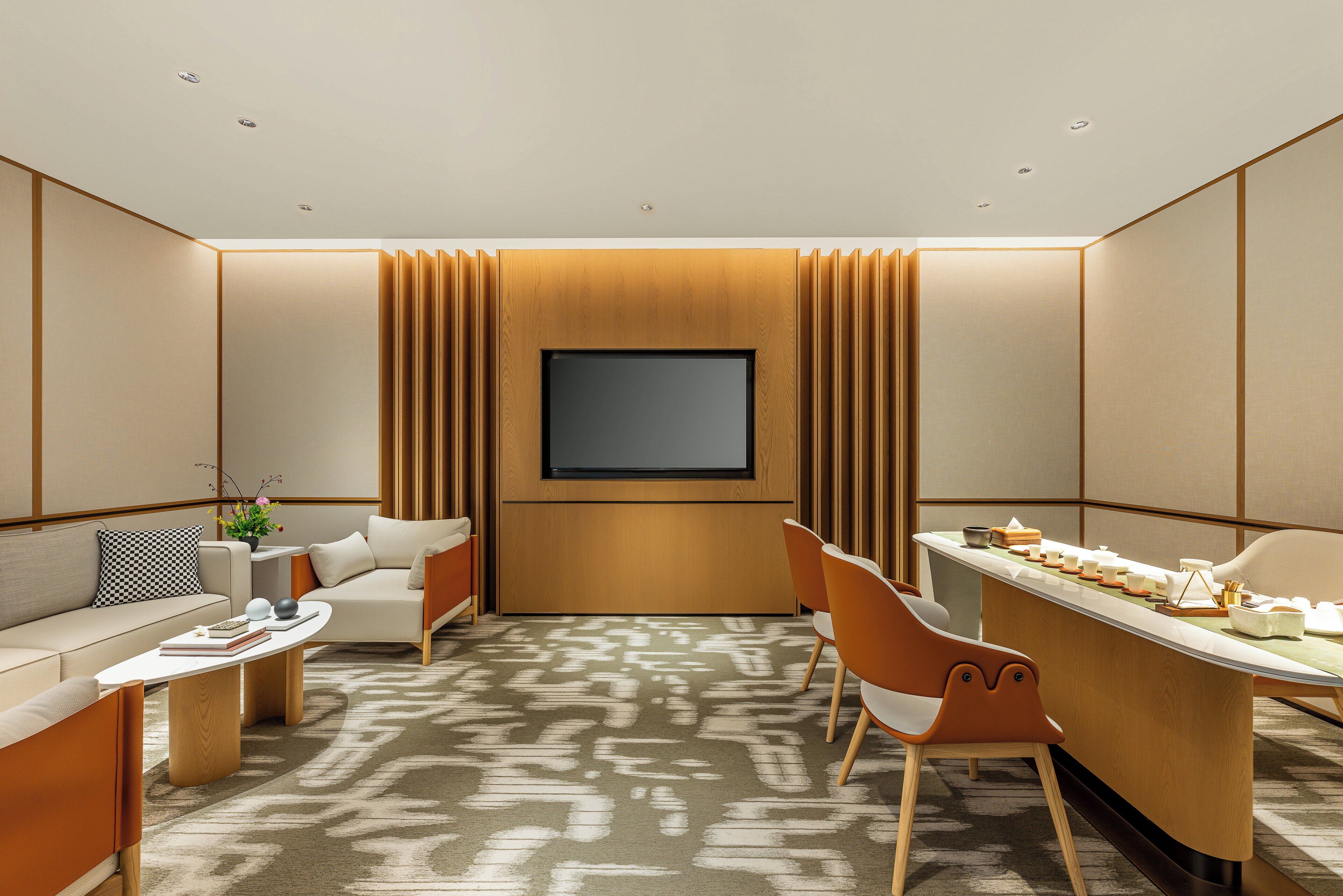 Una habitación de hotel moderna con un televisor de pantalla plana montado en la pared, un sofá, una mesa de comedor con sillas y una alfombra estampada.