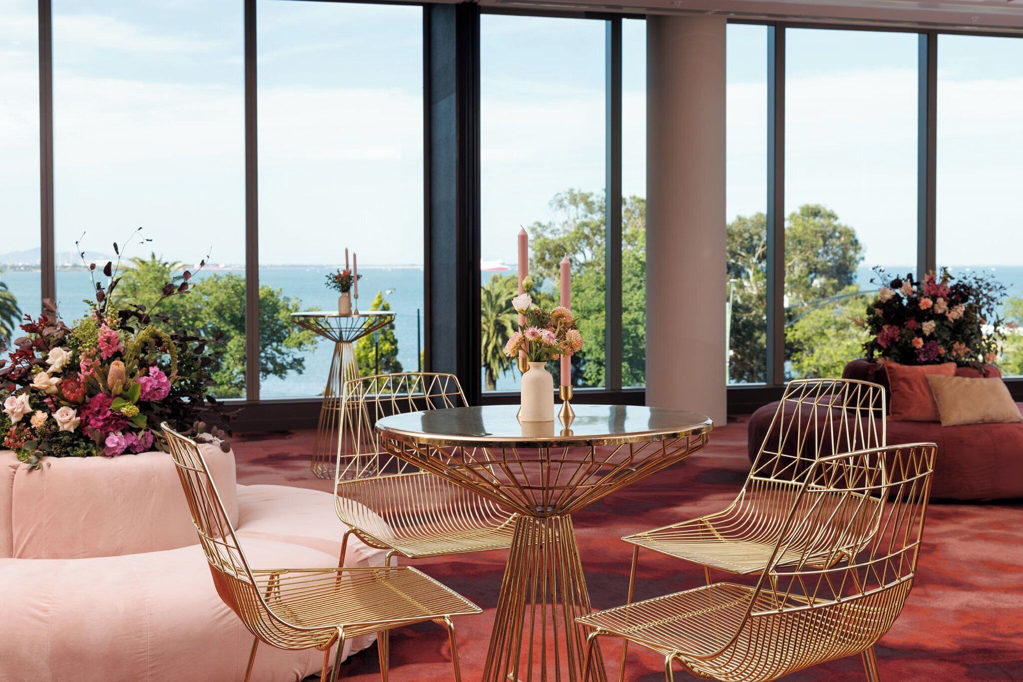 A room with a round table, four chairs, and a sofa. There are large windows with a view of the sea and trees.