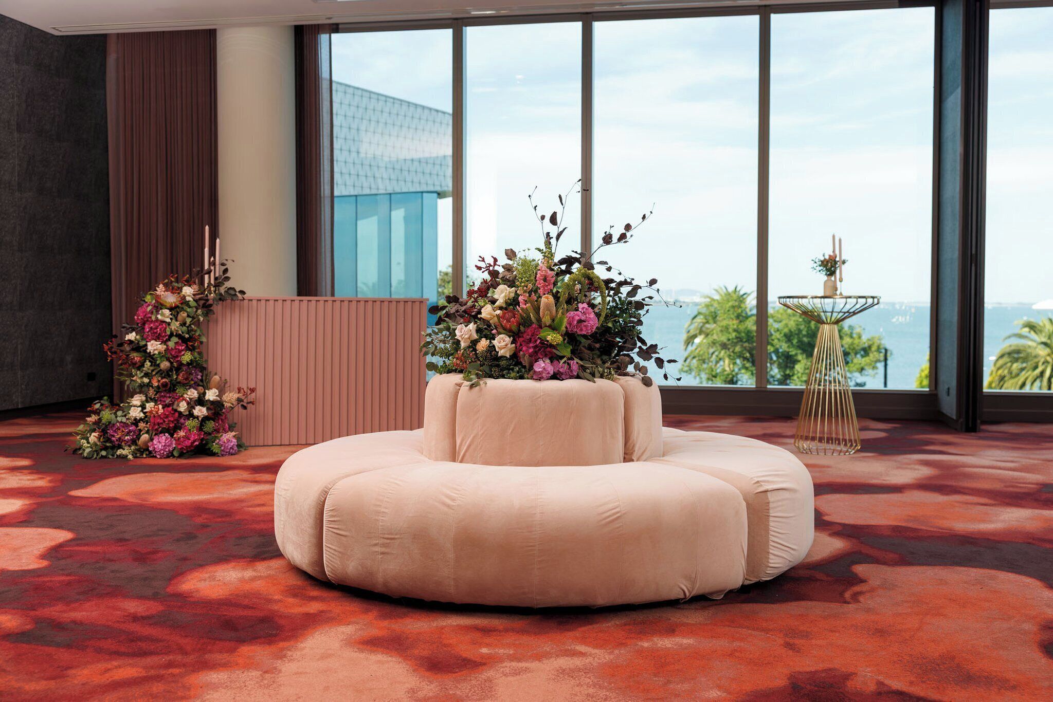 A spacious room with a large window offering a view of the city skyline. A round, cream-colored sofa is positioned in the center, flanked by two floral arrangements.