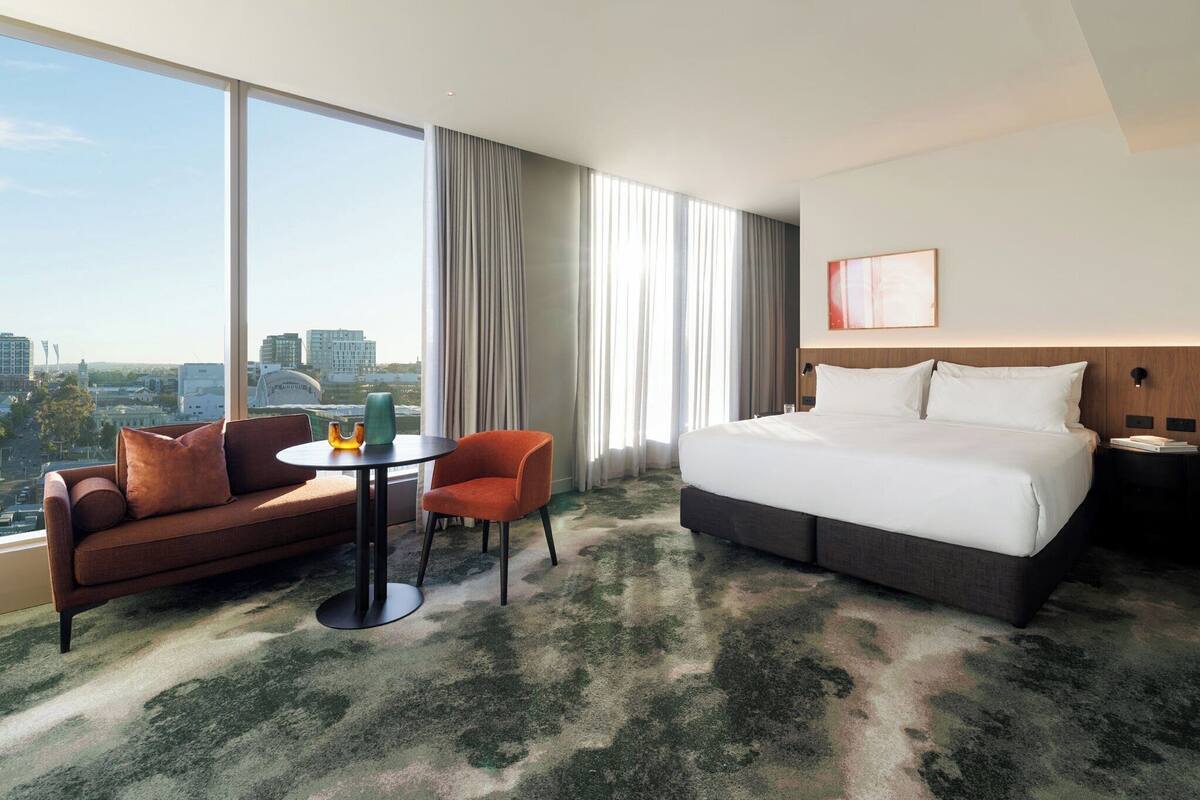Une chambre d’hôtel avec un grand lit, un canapé, une petite table et une chaise. Il y a une grande fenêtre avec des rideaux, et on peut admirer la vue sur la ville depuis l’extérieur.