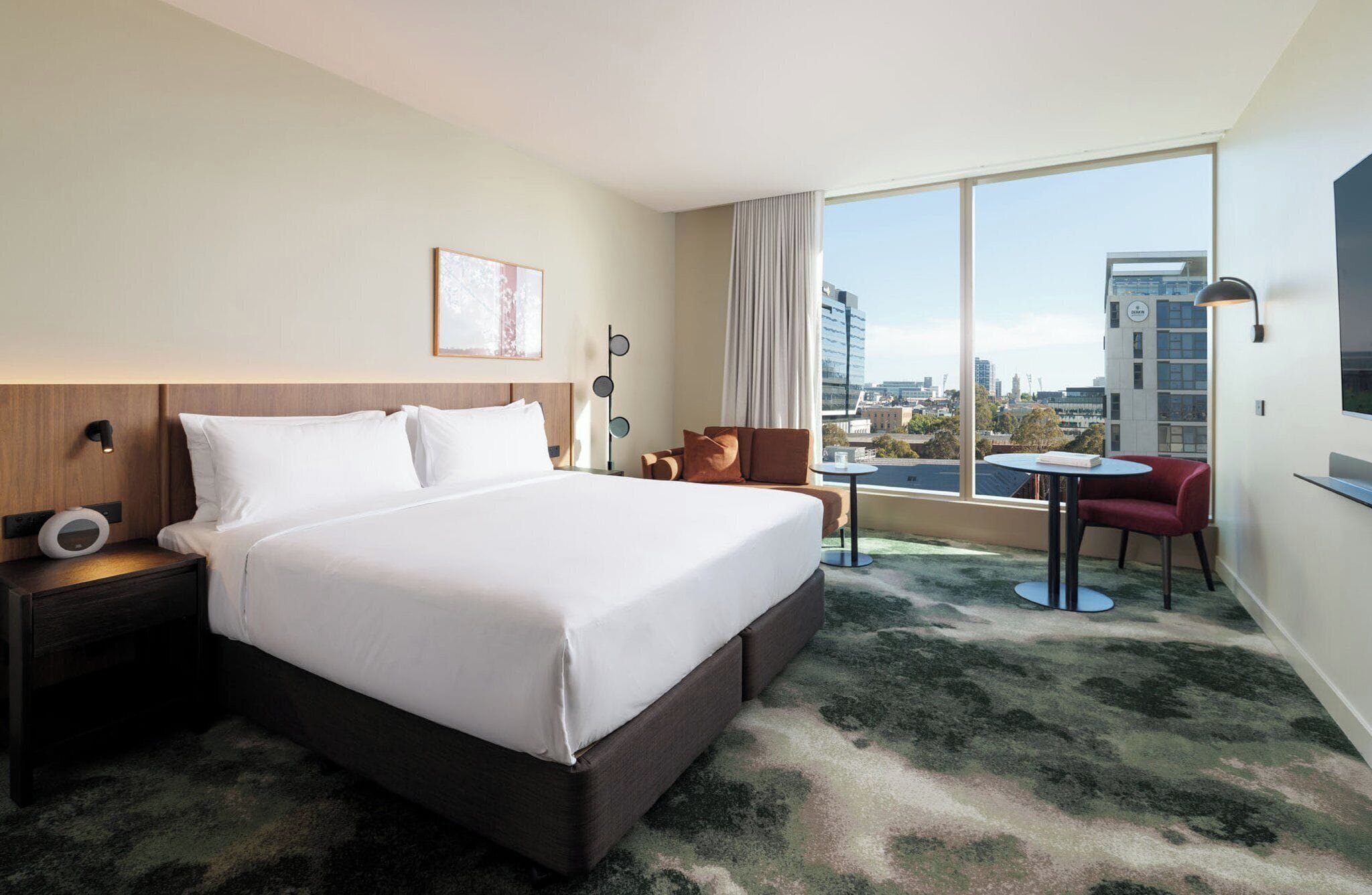 Une chambre d’hôtel avec un grand lit, une petite table, une chaise rouge, une vue sur la ville et une grande fenêtre.