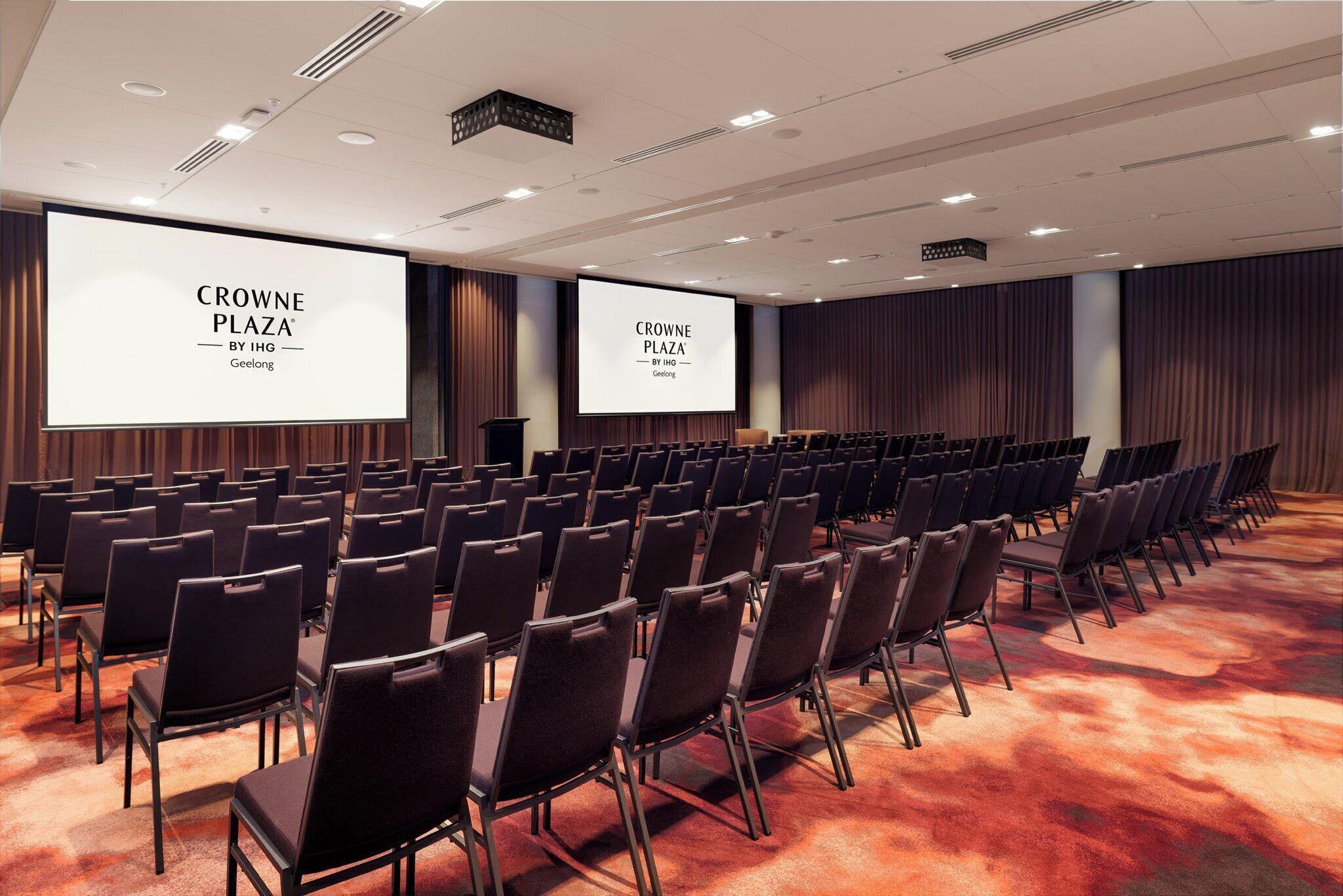 Uma sala de conferências com fileiras de cadeiras e uma tela grande exibindo 'CROWNE PLAZA BY IHG Geelong'.