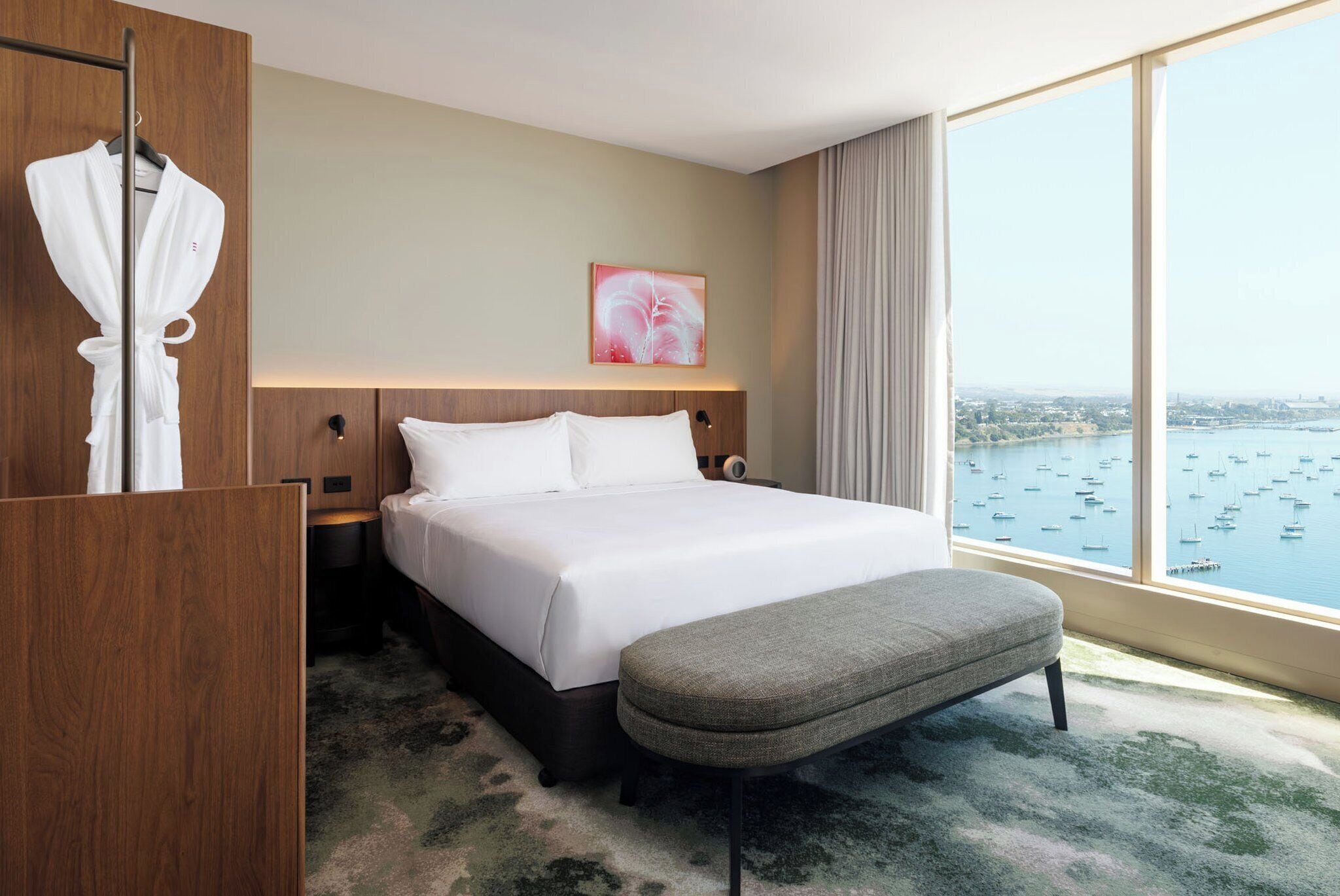 A hotel room with a large bed, a grey bench, a robe rack, and a city view through large windows.