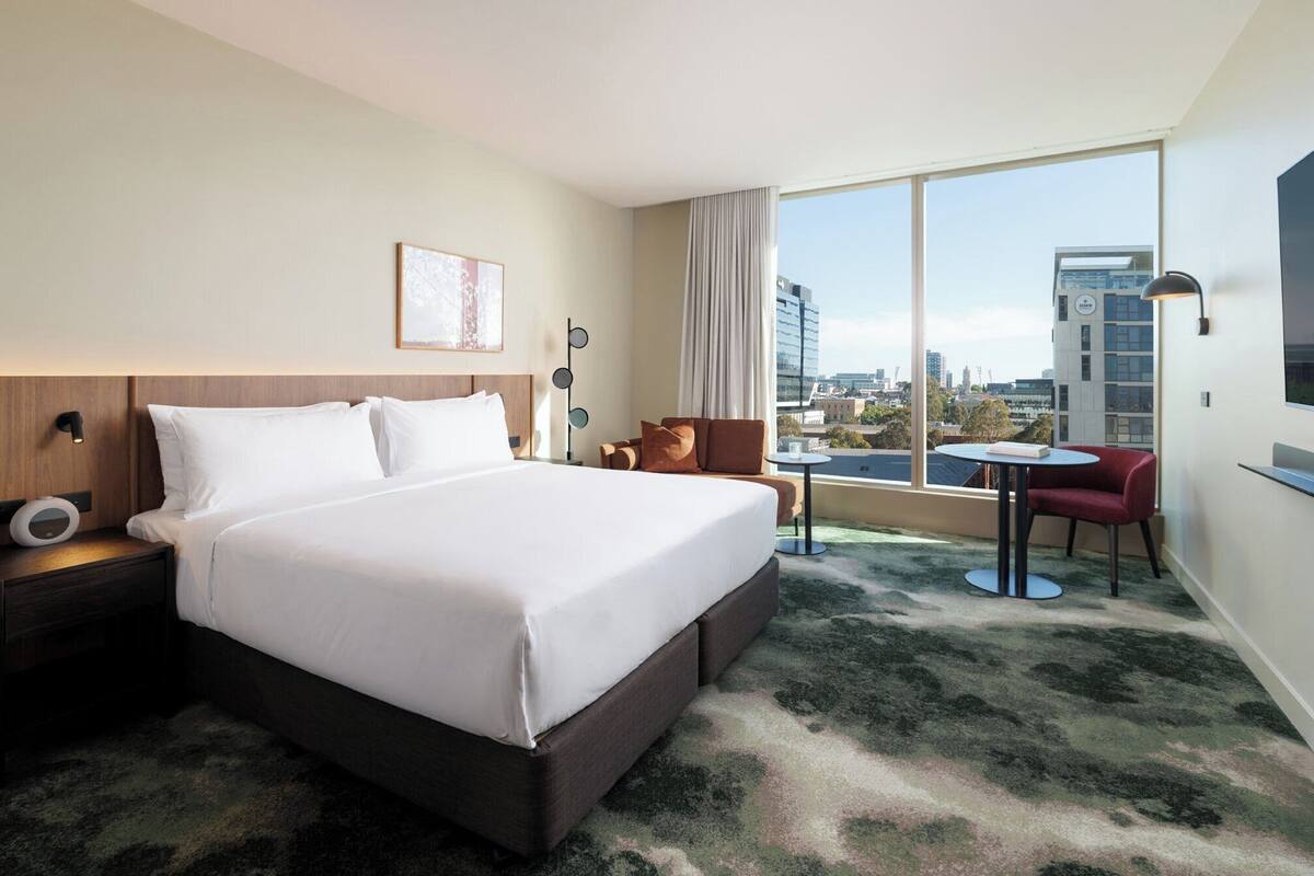 Une chambre d’hôtel avec un grand lit, une petite table, une chaise rouge, une vue sur la ville et une grande fenêtre.