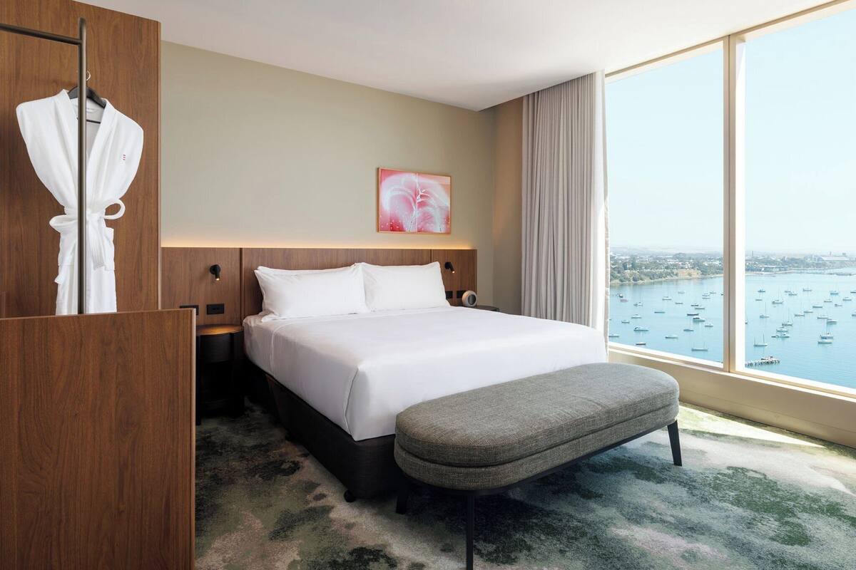 Une chambre d’hôtel avec un grand lit, un banc gris, un porte-peignoir et une vue sur la ville à travers de grandes fenêtres.