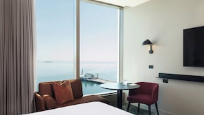 Una habitación de hotel con un ventanal grande, una cama, un sillón rojo, una mesita redonda y un televisor de pared.