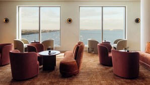 Una habitación de hotel moderna con ventanas amplias, diversas opciones de asientos y vista al mar.