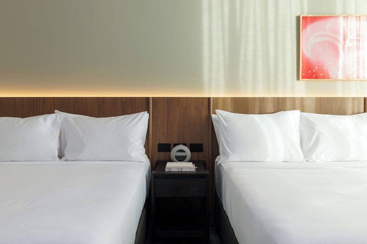 Habitación de hotel con dos camas, cabecero de madera, mesita de noche con lámpara y una obra de arte abstracta colgada en la pared.