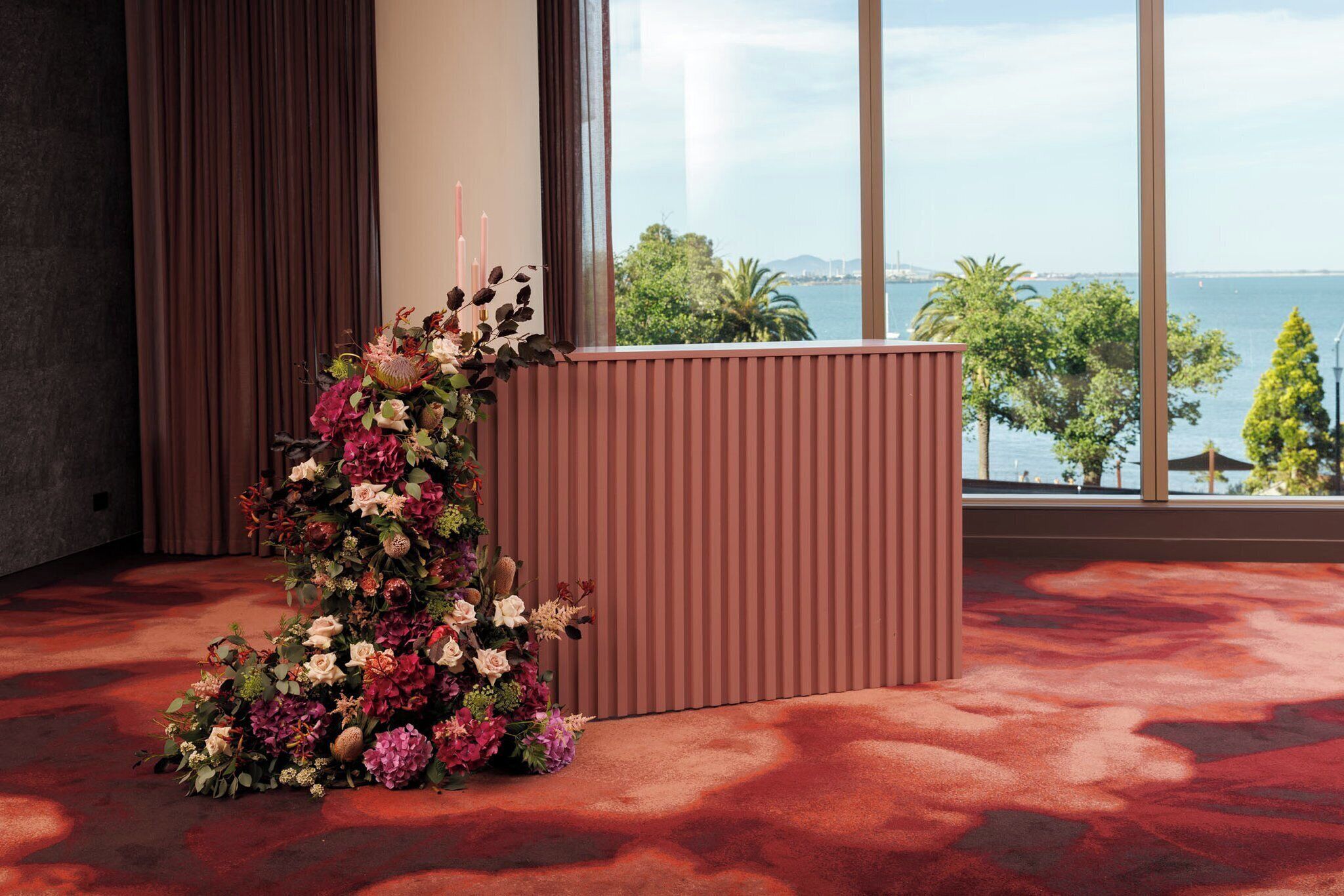 A room with a large window, a red carpet, and a floral arrangement on a podium.
