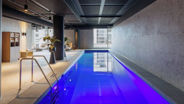 Una piscina interior con fuente de agua iluminada en azul, diseño de interiores moderno con techos a la vista y vistas de la ciudad.