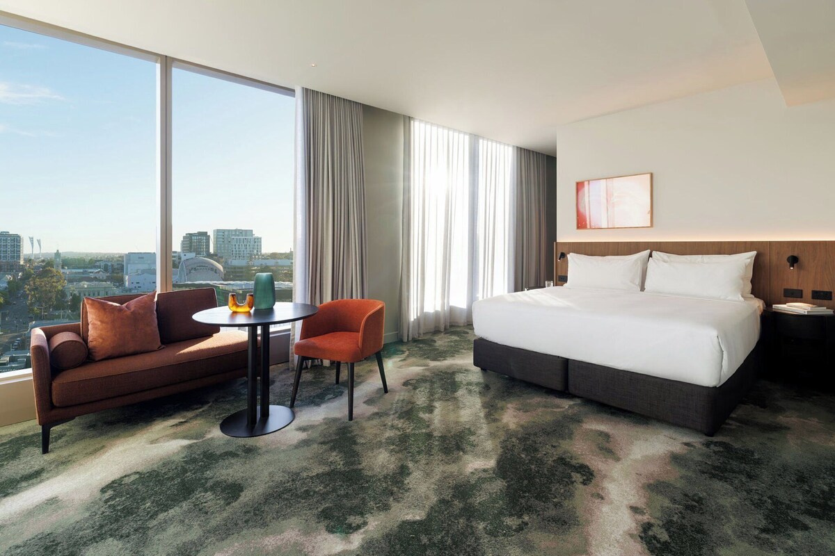 Habitación de hotel con una cama grande, un sofá, una mesita y una silla. Tiene un ventanal con cortinas y se ve una vista de la ciudad.