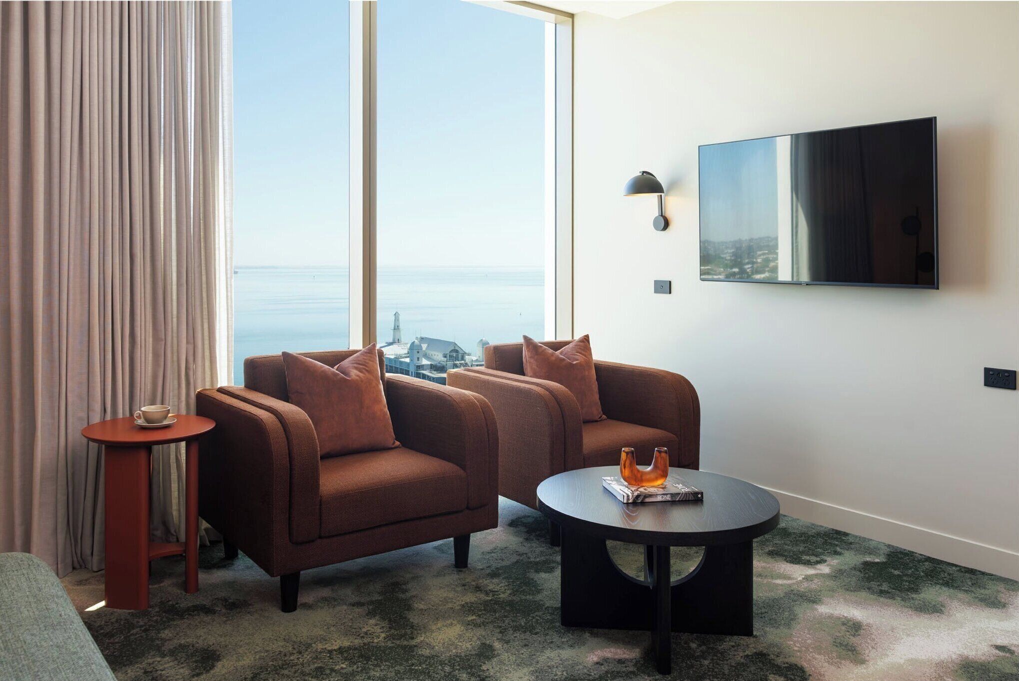 A modern hotel room with a large window offering a city view, a flat-screen TV mounted on the wall, a round coffee table, and two brown armchairs.