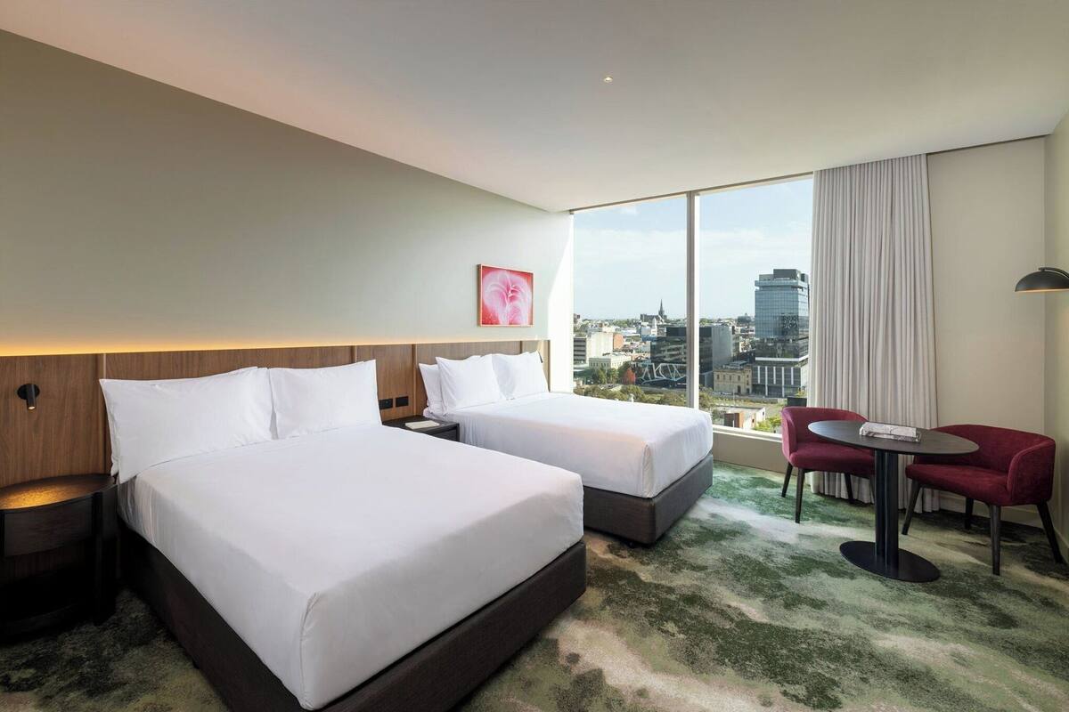 Une chambre d’hôtel avec deux lits, une vue sur la ville, un fauteuil rouge et une petite table.